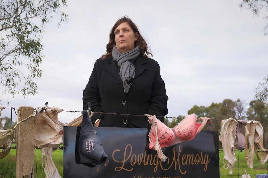Close up of a woman behind a sign reading 'In loving memory of those forever in our hearts' with some bras hanging either side.