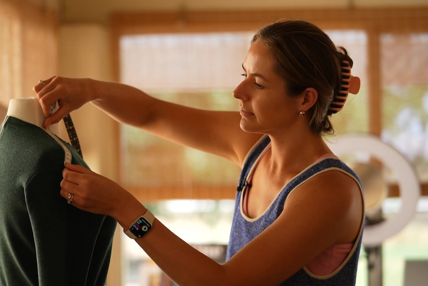 Photo of a woman in a blue singlet with brown hair tied back measuring clothing on a mannequin. 