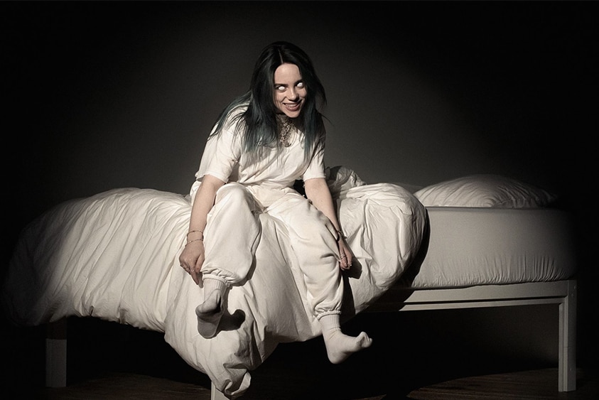 Billie Eilish, dressed in white, sitting down on the edge of the bed