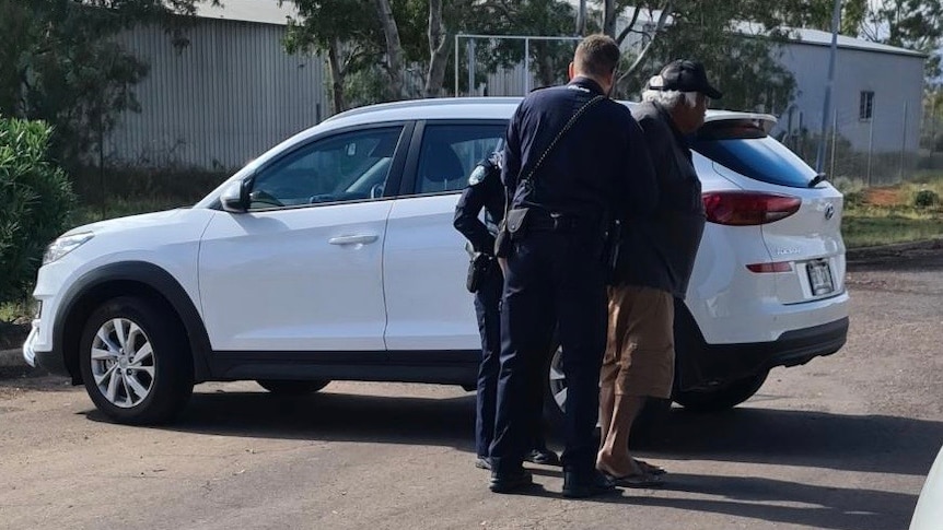 An Aboriginal man is arrested by two police officers. A white car is in the background.