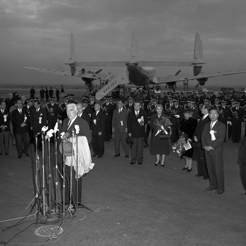 black and white image of menzies in front of many microphones at an airport with crowd of officials standing behind him