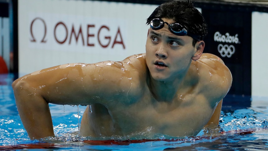 Swimmer Joseph Schooling is in a pool, pulling his body up over a lane rope to look up at a scoreboard. 