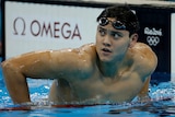 Swimmer Joseph Schooling is in a pool, pulling his body up over a lane rope to look up at a scoreboard. 