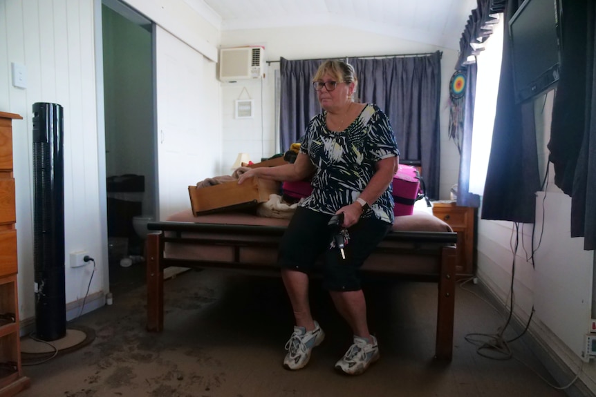 A woman wearing glasses sits on a bed with drawers and other belongings, the floor is covered in mud.