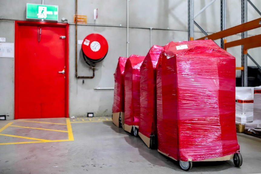 Large rectangular boxes on wheels wrapped in red-plastic in a row next to an emergency exit door on a warehouse floor