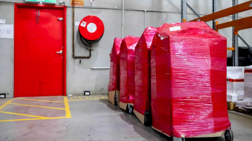 Large rectangular boxes on wheels wrapped in red-plastic in a row next to an emergency exit door on a warehouse floor