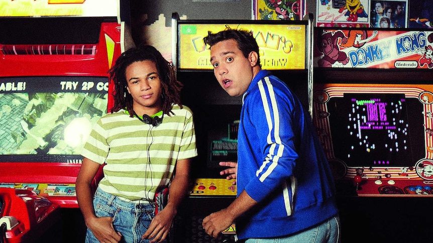 Kamil Ellis (left) and Calen Tassone (right) stand in front of three old video game cabinets.