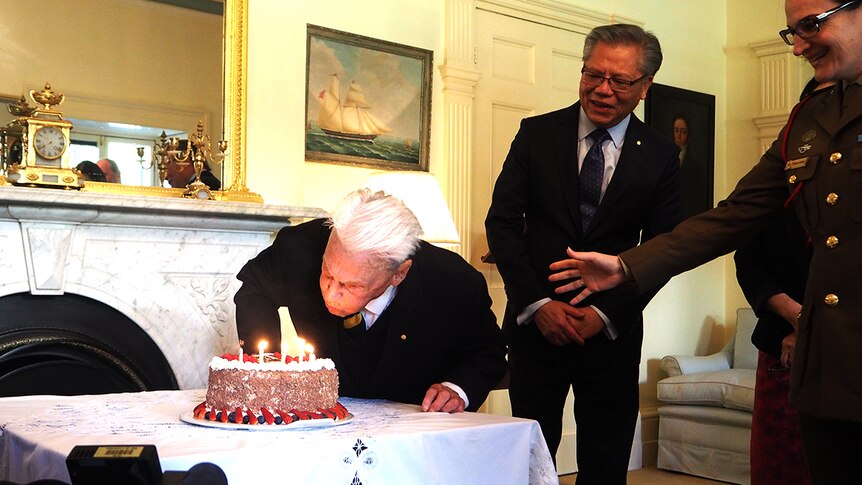An elderly man blows out a candle on a cake while others watch on.