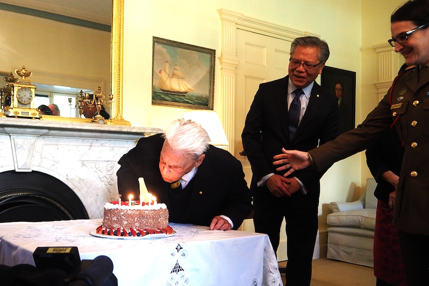 An elderly man blows out a candle on a cake while others watch on.