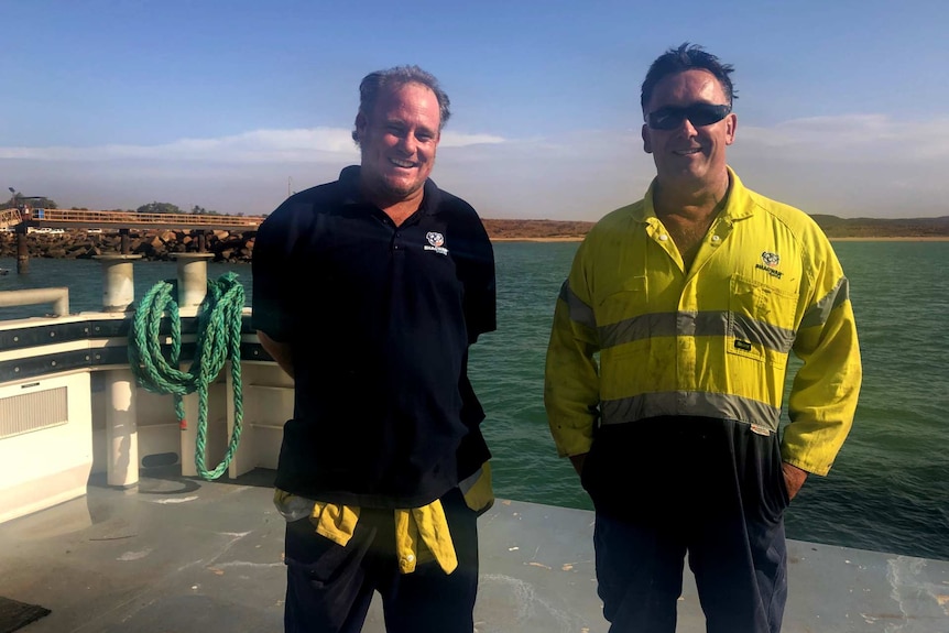 Two men in work clothes smiling at camera, standing beside the water.