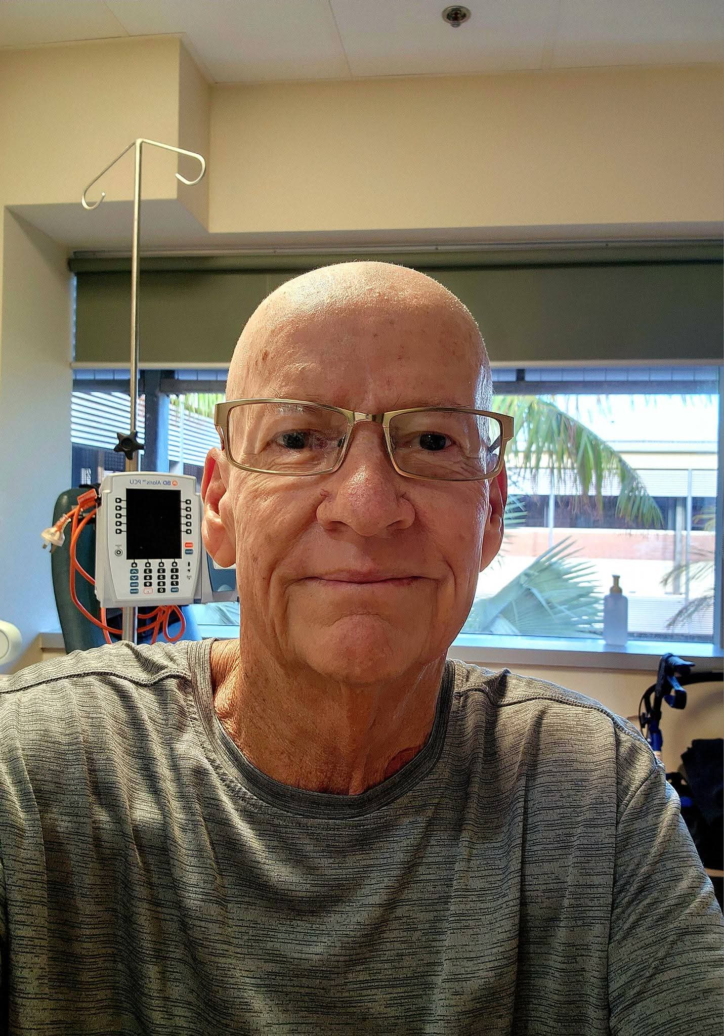 A bald man wearing reading glasses and a grey shirt looks striaght ahead