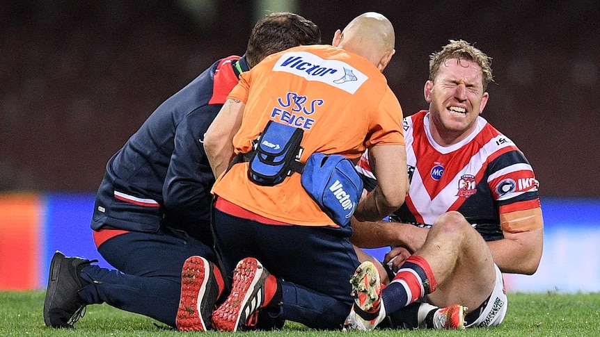 Mitch Aubusson grimaces on the ground and holds his arm as two men kneel next to him