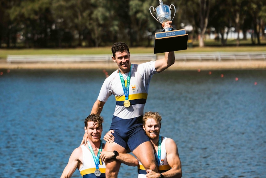 Two male rowers carry another male rower on their shoulders. The rower on the shoulders is lifting a trophy. 