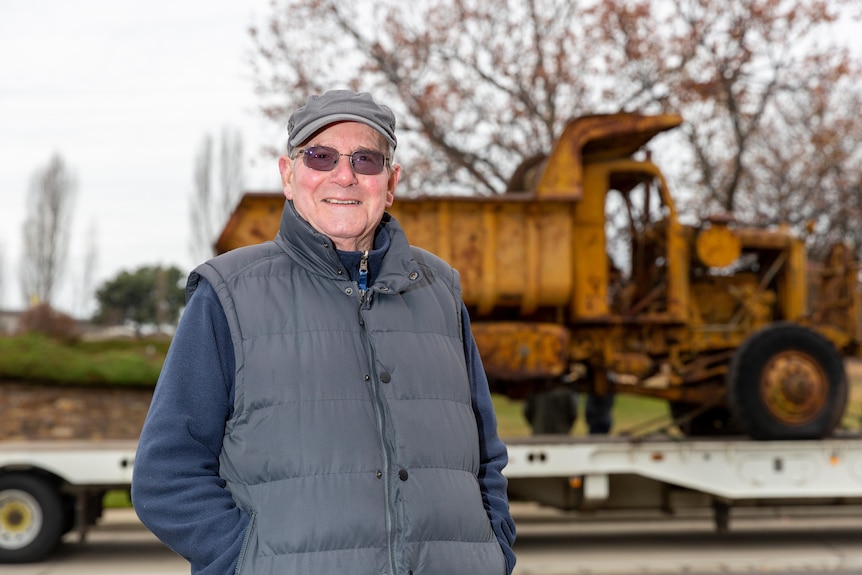 Older man wearing navy fleece top, grey vest and cap stands in front of truck smiling at camera.