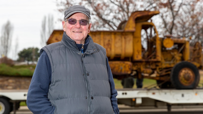 Older man wearing navy fleece top, grey vest and cap stands in front of truck smiling at camera.
