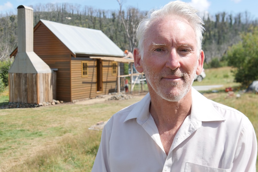 close up of man with white hair with a wooden hut in the background