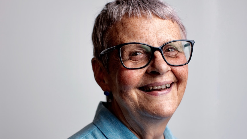 Wendy Mitchell with black-framed glasses, short grey hair and blue shirt turns slightly and smiles widely towards the camera.
