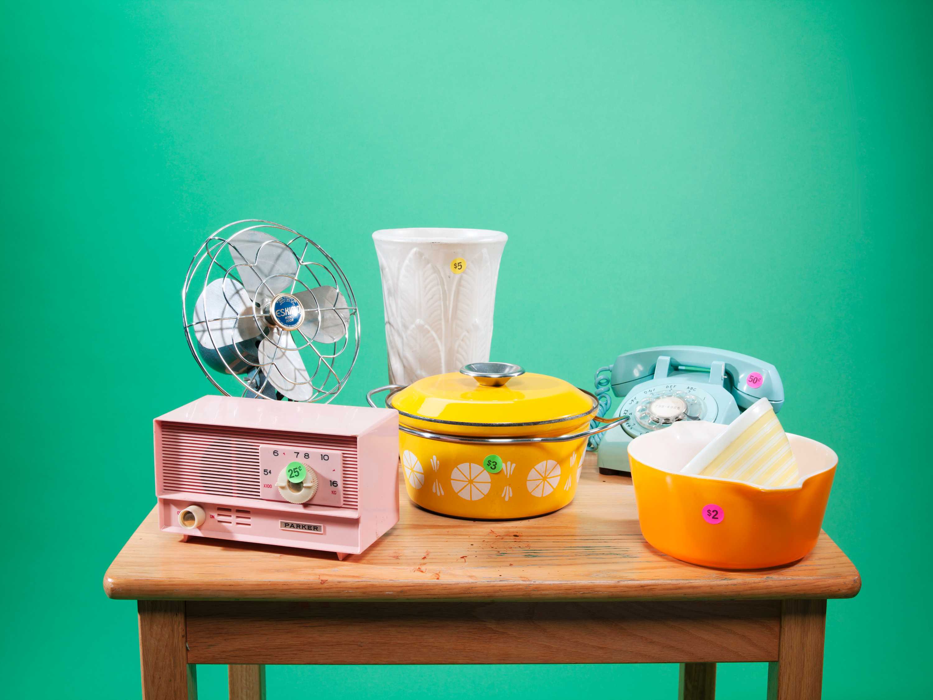 A fan, vase, radio, rotary phone and tableware are displayed on a wooden table with low-price stickers.