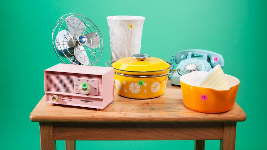 A fan, vase, radio, rotary phone and tableware are displayed on a wooden table with low-price stickers.