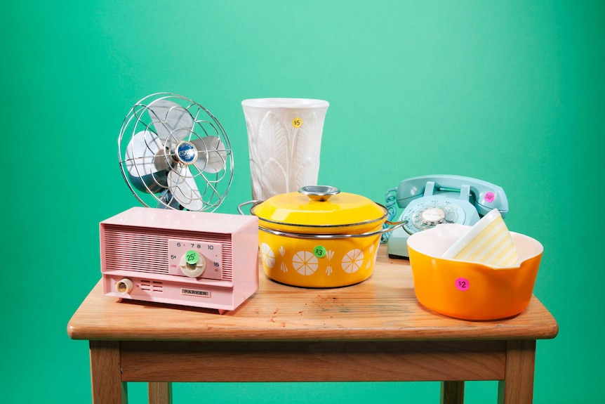 Un ventilateur, un vase, une radio, un téléphone à cadran et de la vaisselle sont exposés sur une table en bois avec des autocollants à petit prix.