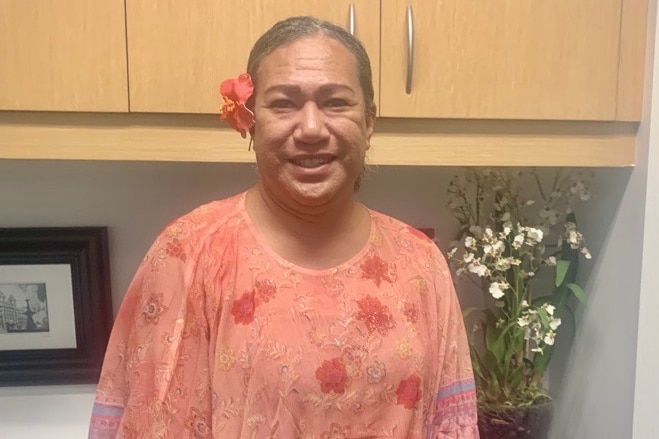 A samoan woman wearing a flower in her ear smiles in a small kitchen area.