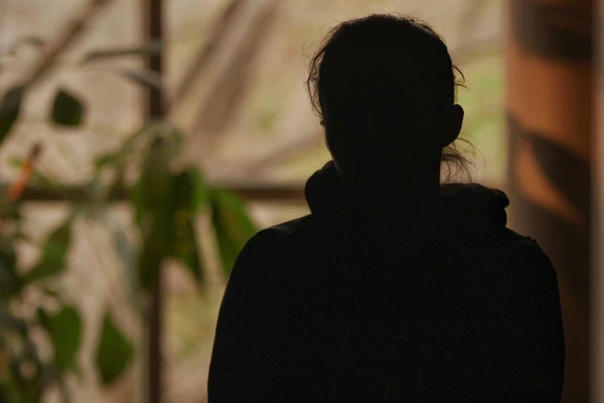 An unidentified woman sits in the shadows of a building foyer with plants behind her.