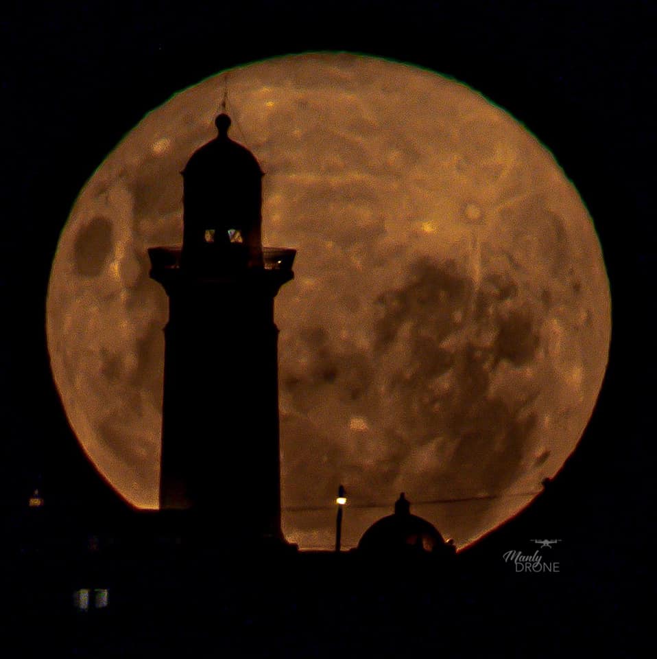 The moon behind the black silhouette of a lighthouse. 