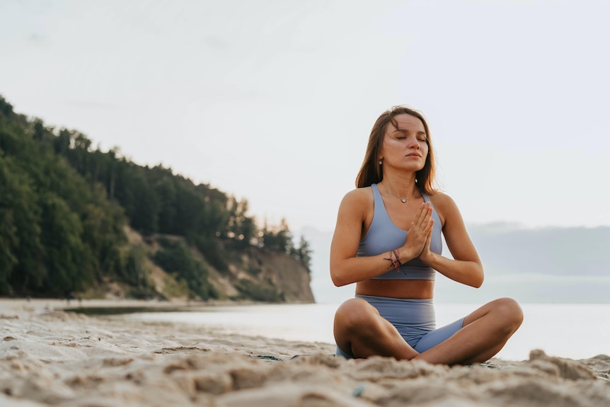 A woman in blue fitness wear sits cross-legged on a beach, palms pressed together and eyes closed.