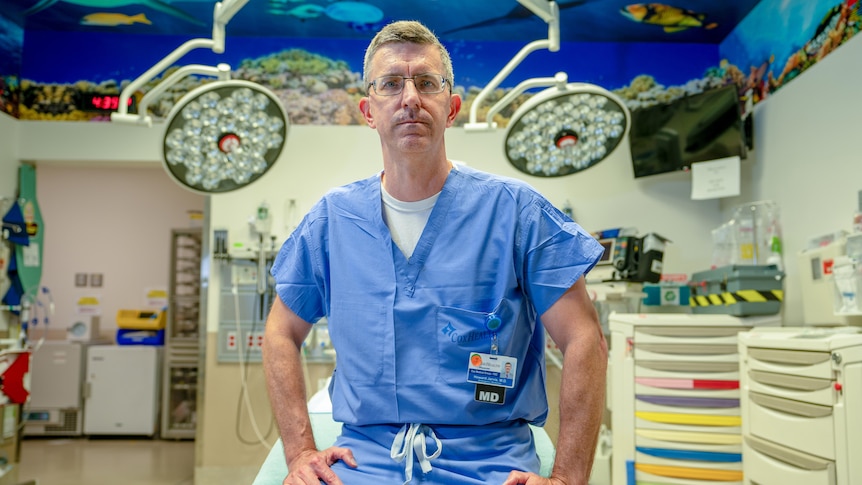 A man in glasses and blue scrubs sits on a hospital operating table 