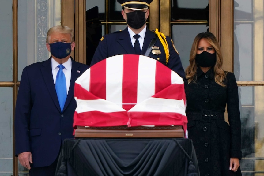 Donald Trump and Melania Trump in face masks standing on either side of a coffin draped with the US flag