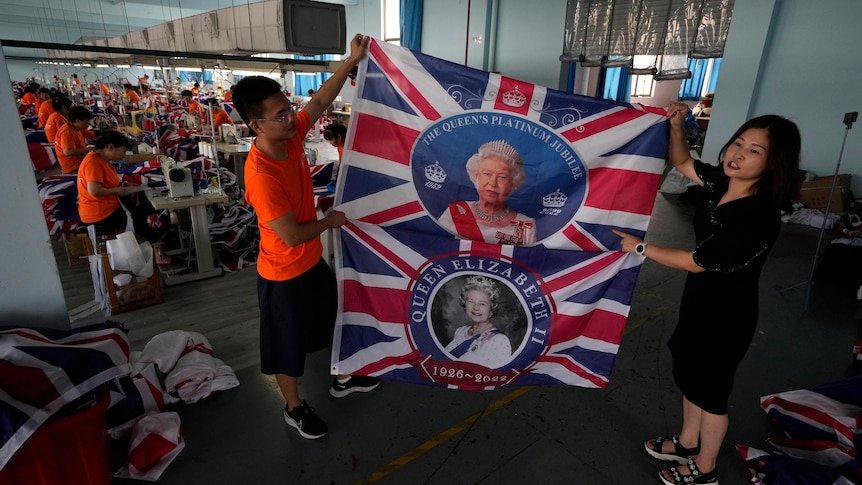 On the right a woman and a man are holding up two British flags with an image of the Queen. In the background workers are sowing