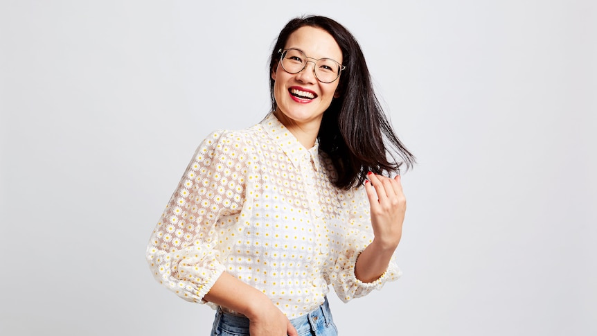 A 30-something Asian Australian woman with glasses smiles broadly while flicking her hair to one side.