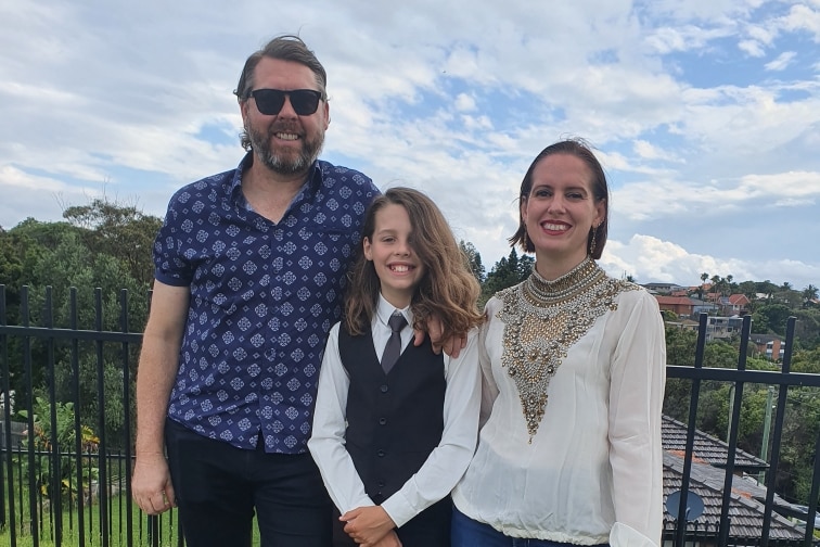 Three people huddled together in front of a fence smiling at the camera