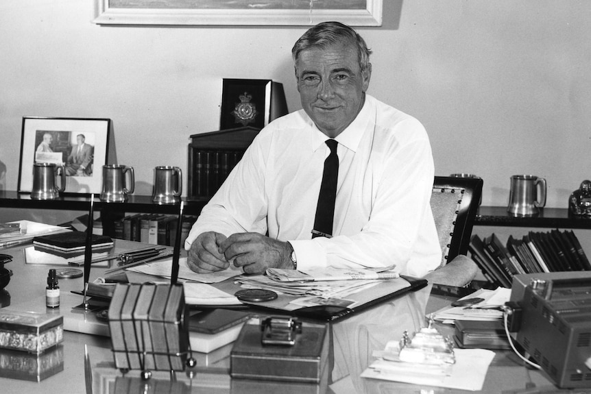 A black and white image of a smiling Charles Moses in a white shirt, seated at his desk with his hands clasped.