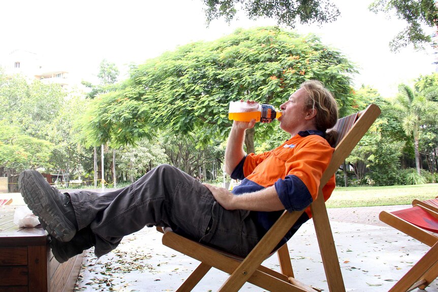 A workman sits in a canvas chair under trees with his feet up while having a drink from a bottle.
