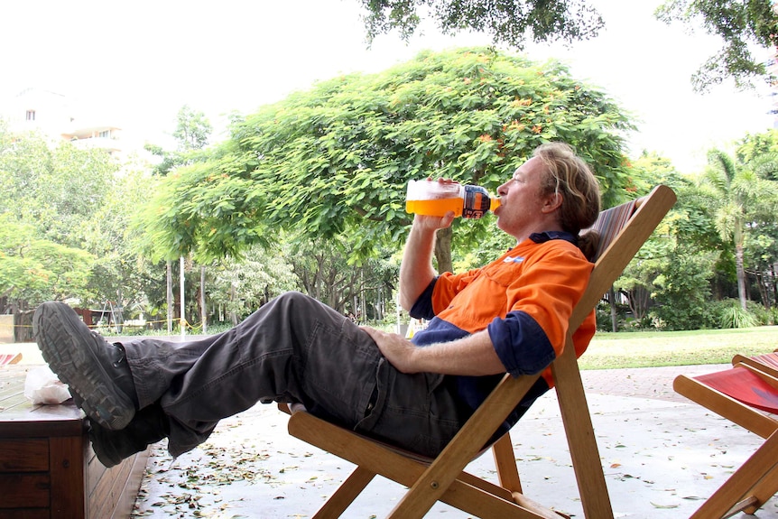 A workman sits in a canvas chair under trees with his feet up while having a drink from a bottle.