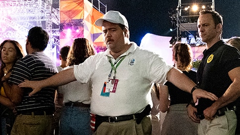 Two men wearing cargo pants and white polo and caps hold back crowd at outdoor night time event in a grass area.