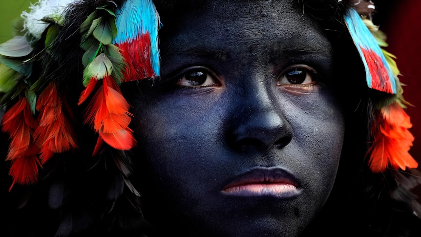 An Indigenous girl with her face dyed all black wearing a colourful feathered headband. 