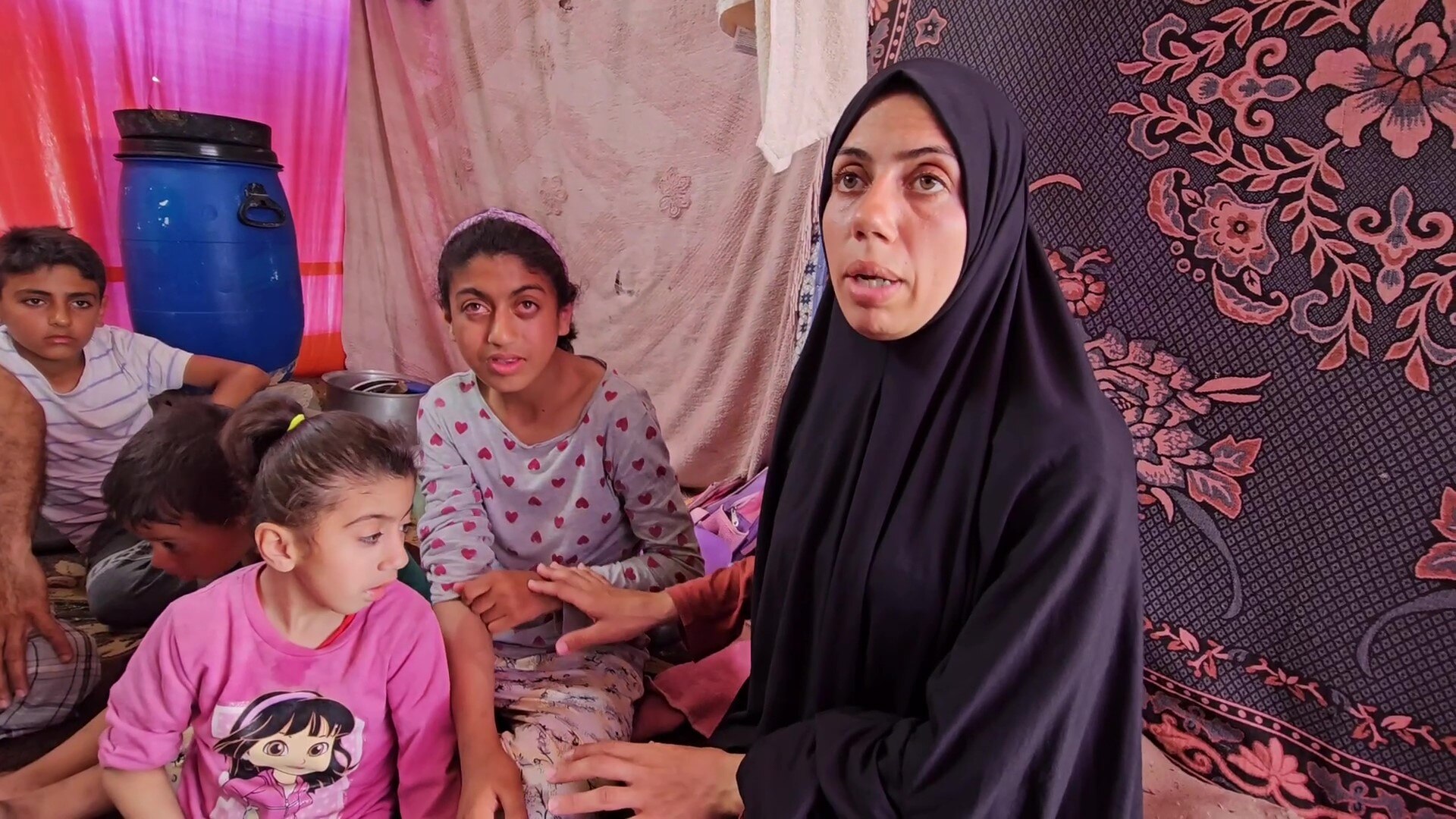 A woman in a black hijab sitting in a tent and pointing at children