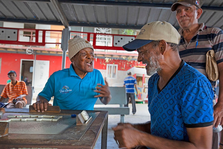 Two men laugh at a table in the park while playing dominoes