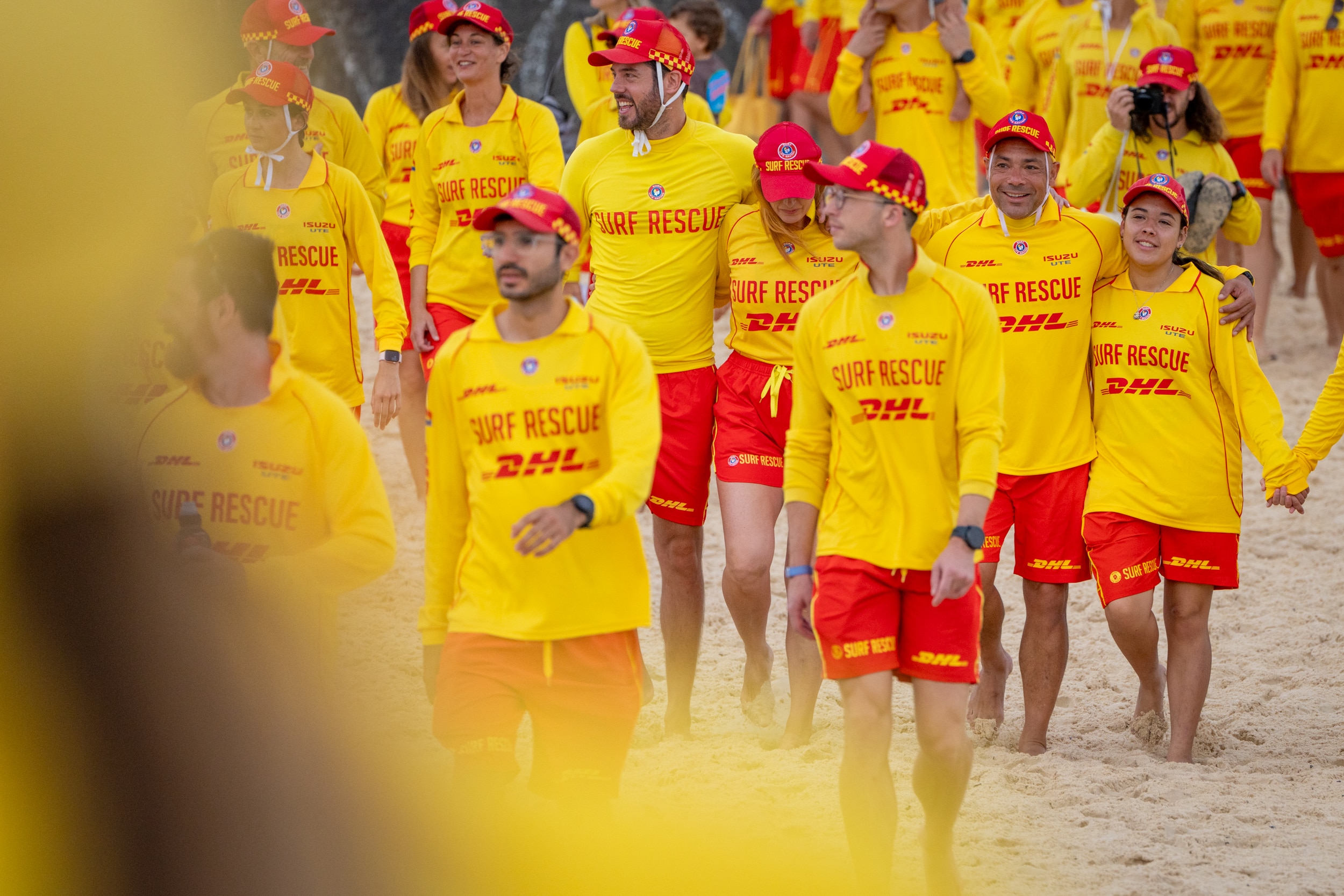 Salva-vidas em uniformes vermelhos e amarelos caminham pela praia, alguns de mãos dadas