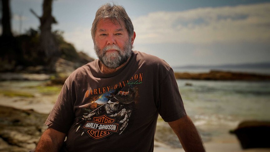 A man sits on a rock at the beach and poses for a photo with a serious expression