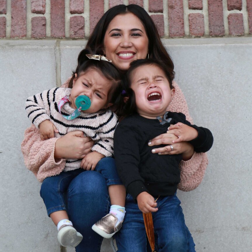 a woman with dark hair crouches down and smiles at the camera, the two children in her arms are losing their shite and crying!