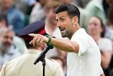 Novak Djokovic stands at a microphone and points his finger at the crowd on Centre Court at Wimbledon.