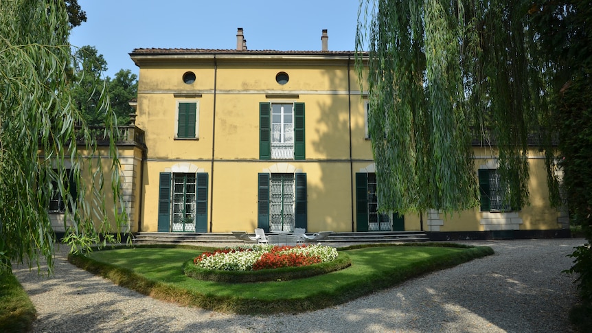 An Italian villa with yellow paint and green shutters, framed by willow trees.