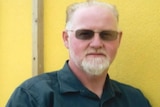 A man with grey hair wearing sunglasses in front of a yellow wall.