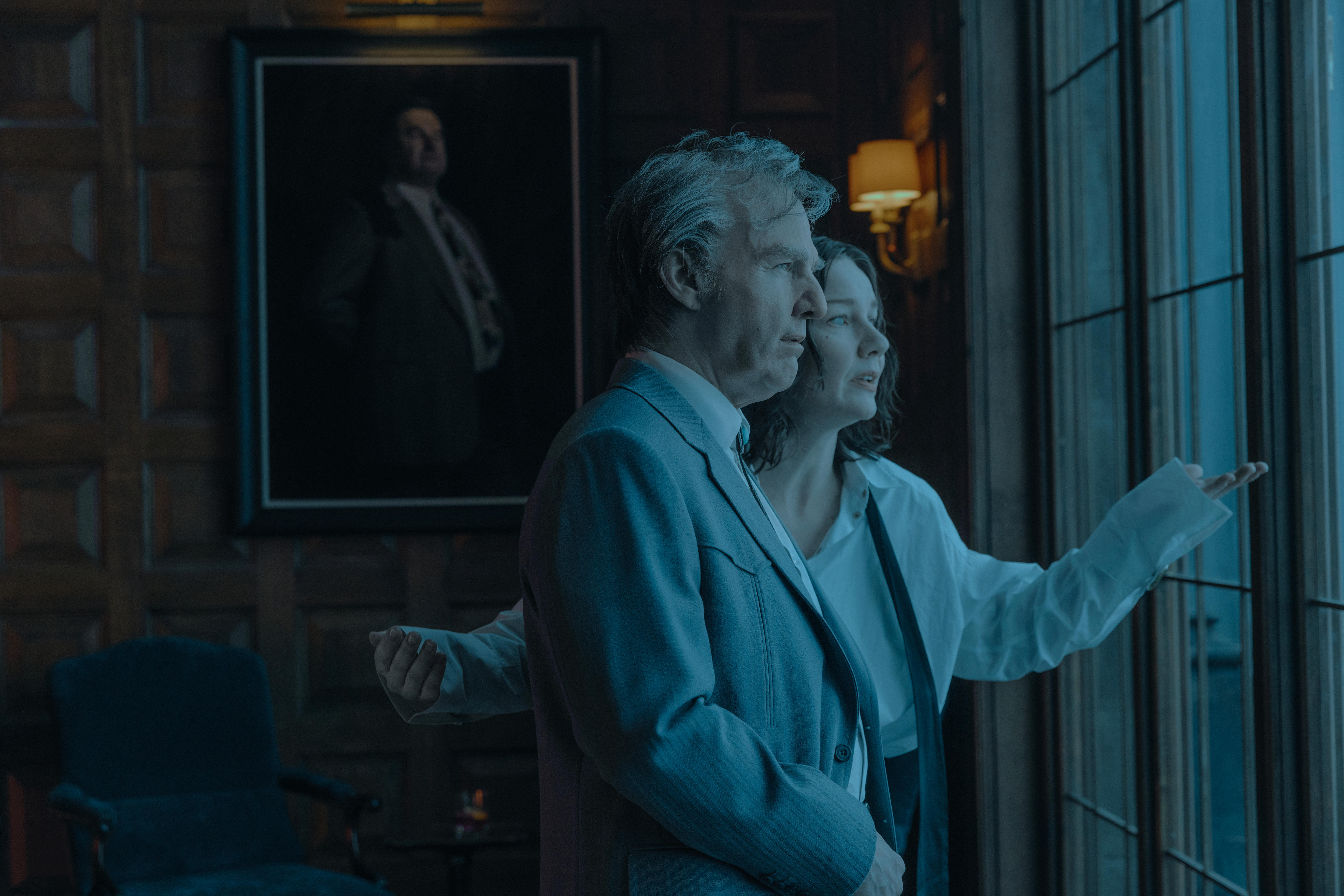 A man and a woman stare out of the large window in a darkened room.