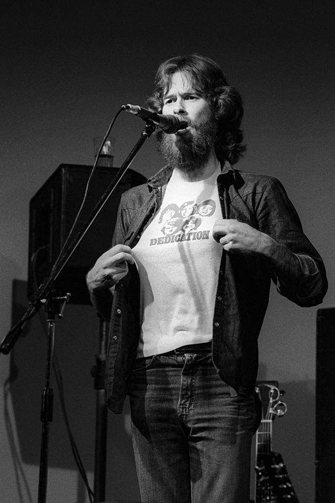 A man with flowing long hair stands behind a microphone holding his t-shirt out that says 'dedication'