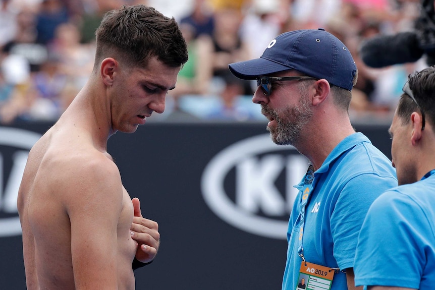 A shirtless Thanasi Kokkinakis presses his pectoral while talking to his coach during a medical timeout on court