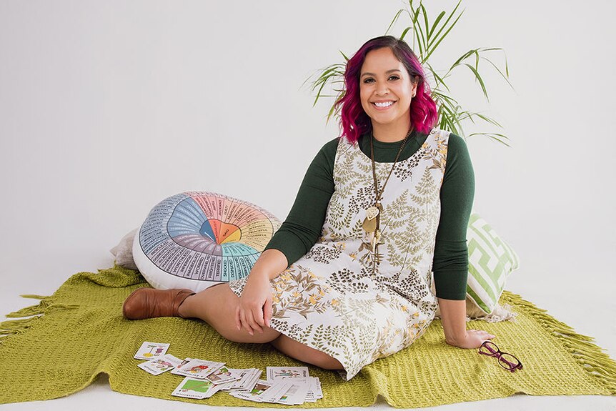 Sarah wears a green shirt and white patterned dress. She has pink hair and sits on a green rug with cards a pillow and a plant